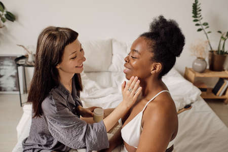 Happy young woman applying facial cream on face of her African girlfriendの写真素材