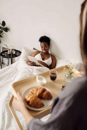 Happy African female with tablet looking at her girlfriend holding tray with breakfastの写真素材