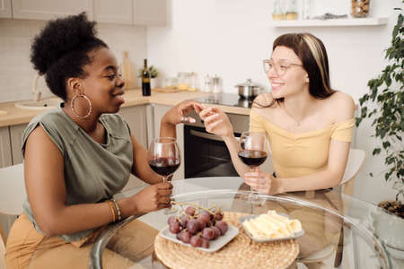 Two happy girlfriends with glasses of red wine sitting by table in the kitchenの写真素材