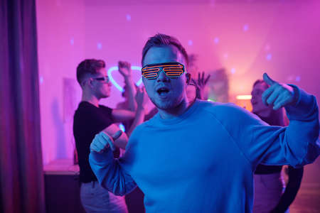 Young serious man in casualwear and disco eyeglasses looking at you while standing in front of his...