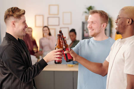 Happy young intercultural men clinking with bottles of beer and looking at one another with toothy smiles while cheering up at home partyの写真素材