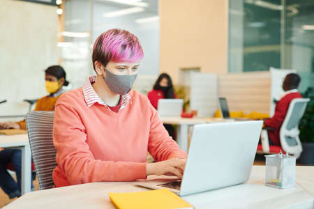 Young female office manager in casualwear and protective mask looking at laptop display while preparing presentation against her colleaguesの写真素材