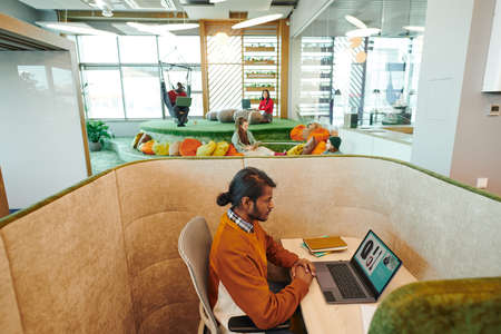 Young serious mixed-race businessman touching keyboard buttons while sitting by desk in front of...