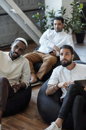 Three young Muslim businessmen sitting in armchairs and listening to speaker or coachの写真素材