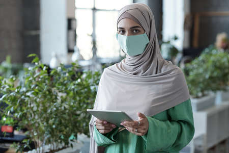 Young Muslim businesswoman in hijab and protective mask looking at camera