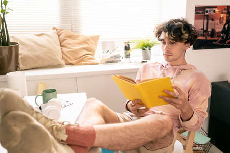 Restful teenager sitting on chair and reading book with his legs on desk