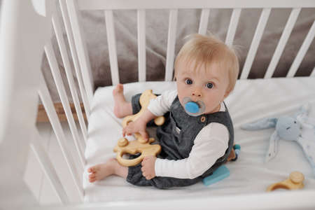 Portrait of serious baby boy with dummy sitting in bed and playing with wooden toy