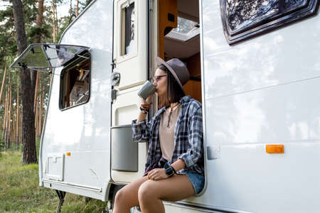 Young woman in hat and casualwear drinking tea while relaxing in house on wheels on hikeの写真素材