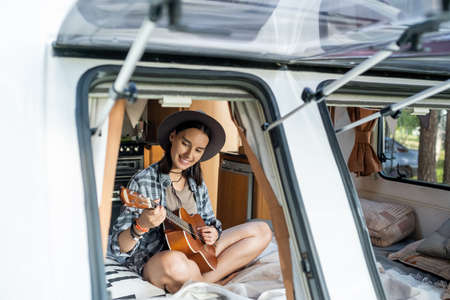 Smiling young woman playing guitar while sitting on bed in small house on wheels
