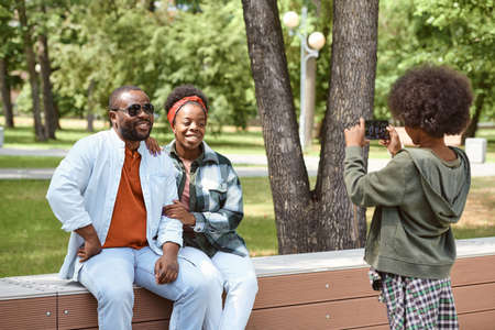 Little African boy with smartphone photographing his happy parents in parkの写真素材