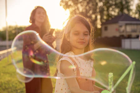 Cute little girl playing with soap bubbles outdoor against her happy motherの写真素材