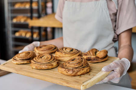 Gloved female baker carrying wooden tray with fresh appetizing buns