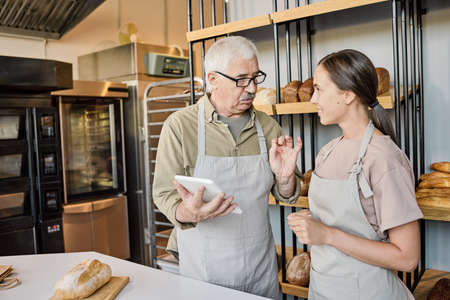 Confident mature baker with tablet consulting with his young female colleague about working pointsの写真素材