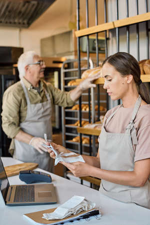 Busy female worker of bakery looking through receits while her mature colleague standing by displayの写真素材