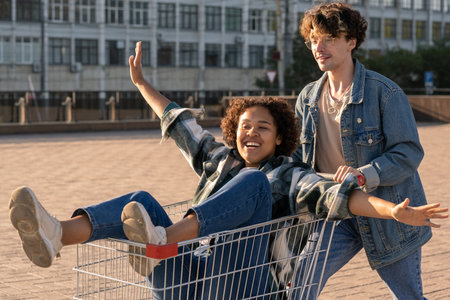 Happy African girl outstretching arms while sitting in shopping cart pushed by her boyfriend