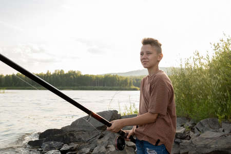 Contemporary teenage boy with long rod fishing alone by lake on summer weekendの写真素材