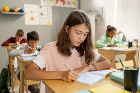 Caucasian elementary schoolgirl making notes while sitting by desk at lessonの写真素材