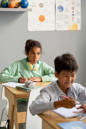 Cute African-American schoolgirl looking at blackboard or teacher at lesson