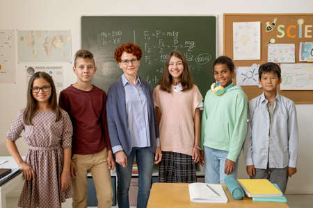Row of happy young teacher and clever schoolchildren standing by blackboard in classroomの写真素材