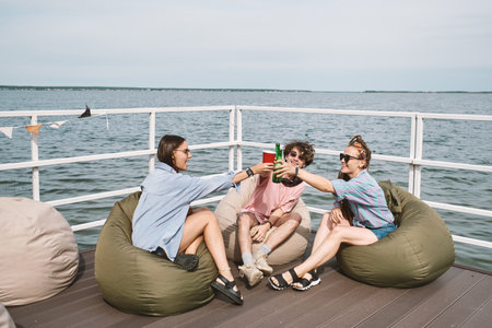 Group of happy young people sitting in bean bags on pier and toasting with beer bottles and plastic cupsの写真素材