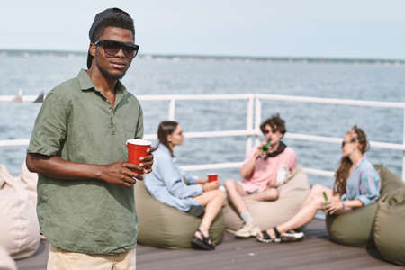 Young man in cap and sunglasses holding red plastic cup, his friend relaxing on bean bags in backgroundの写真素材