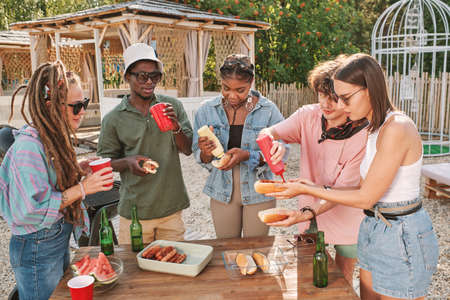 Diverse group of friends drinking beers and eating hot dogs at table on beach enjoying small summer partyの写真素材