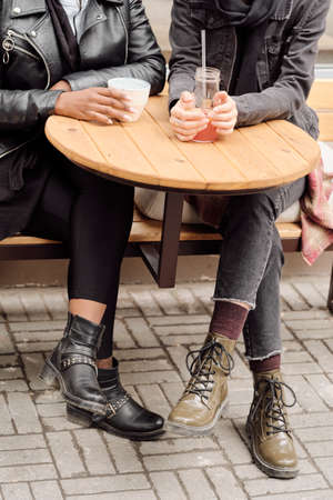Two young intercultural females in casualwear having drinks by round wooden table outdoorsの写真素材