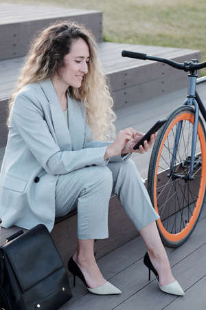Successful young woman in suit scrolling in smartphone while sitting on staircase between bag and...