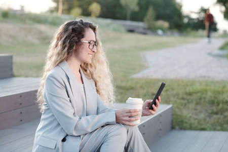 Happy young woman with drink watching video in smartphone while sitting on staircase