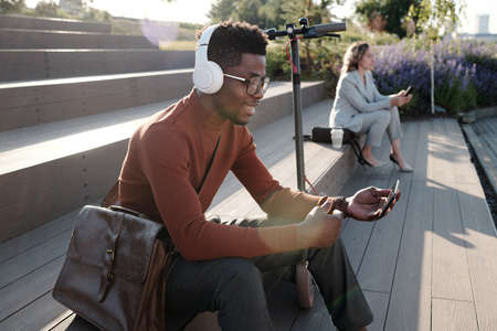 Happy young African man with headphones and smartphone communicating in video chat outdoors