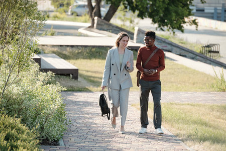 Two happy young businesspeople in smart casualwear moving along public park and interacting