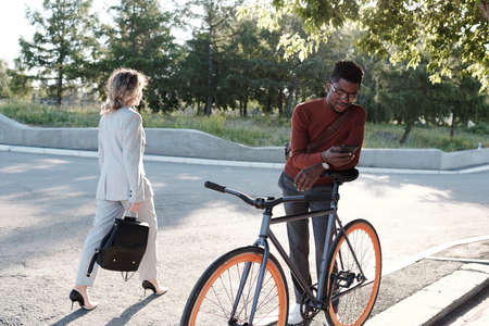 African guy scrolling in smartphone while bending against bicycle with businesswoman passing by...