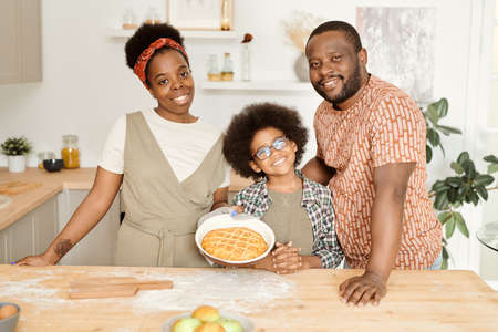 Happy young African family of three with fresh baked apple pie standing by kitchen tableの写真素材