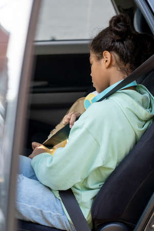 Side view of mixed-race schoolgirl fastening seatbelt while sitting on backseat of car