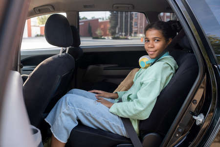 Happy youthful girl looking at you with smile from backseat of car while waiting for parents