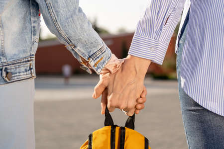Hands of mom and daughter carrying yellow school backpack while moving along street
