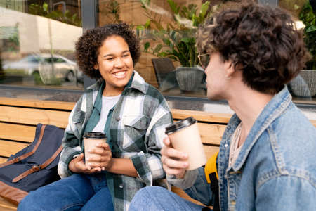 Happy African girl with drink talking to her boyfriend while both sitting on benchの写真素材