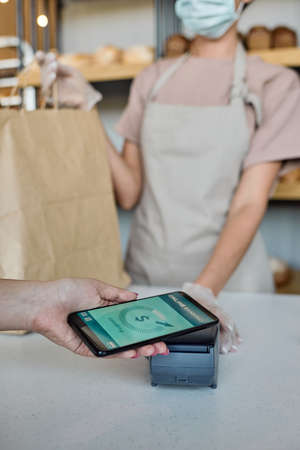 Gloved hand of female baker holding paperbag while customer paying with smartphoneの写真素材