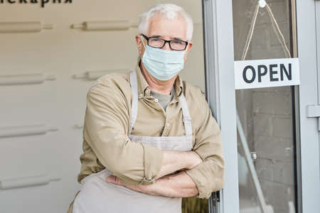 Portrait of senior Caucasian owner of cafe in apron and facial mask standing with crossed arms at...