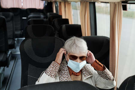 Senior woman in sunglasses sitting in empty bus and putting mask before ride during coronavirus...