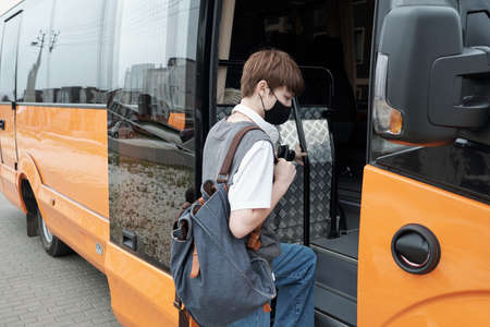 Teenage girl with short hair wearing facial mask holding satchel while getting on contemporary busの写真素材