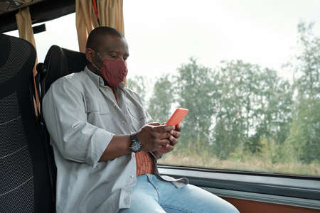 Bald young Black man in mask sitting at window and using smartphone while riding busの写真素材