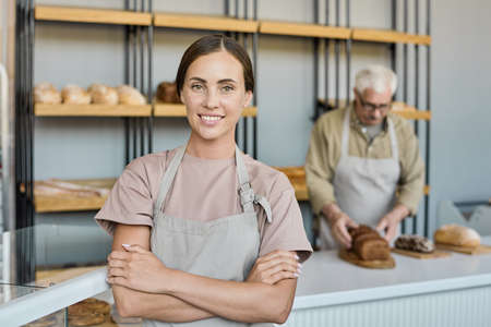Portrait of happy young brunette woman in apron standing with crossed arms in bakery shop owned by...