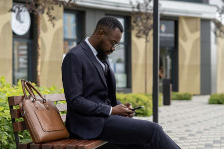 Young businessman of African ethnicity texting in smartphone while resting on bench in urban environmentの写真素材