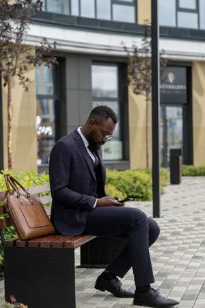 Serious African businessman in suit scrolling in smartphone while sitting on benchの写真素材