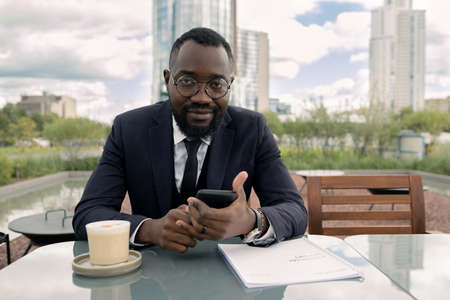 Contemporary young elegant businessman with smartphone sitting by table in outdoor cafe