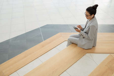 Young Asian female in elegant suit listening to audio in smartphone while sitting on large stairs