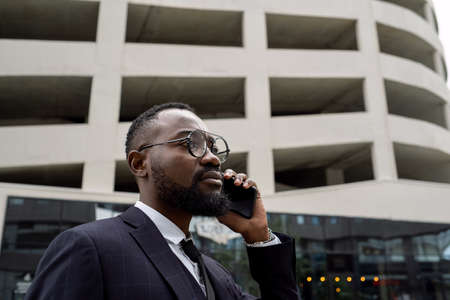 Serious young African businessman holding smartphone by ear while standing against building exteriorの写真素材