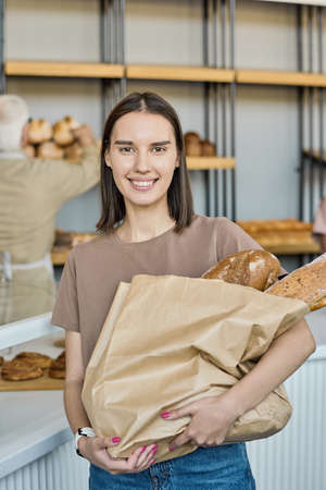 Portrait of happy young female customer standing with paperbag full of loaves of bread in bakery storeの写真素材