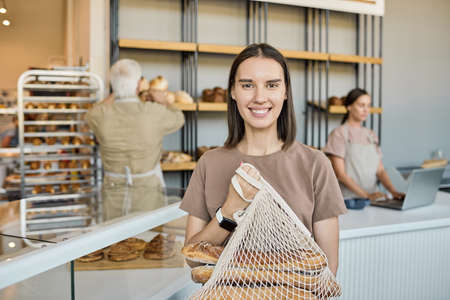 Portrait of happy young female customer holding net bag with fresh loaves of bread against bakery...